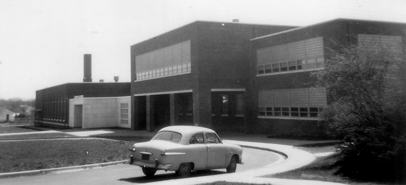  Black and white photograph of the new Herndon High School addition in 1954 from the Fairfax County School Board’s fire insurance survey. It is a long, rectangular, brick building with windows on all sides. A small 1940s era car is parked in the driveway circle in front of the school.