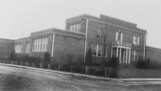 Black and white photograph of 1927 Herndon High School Building, believed to have been taken in March 1937 by a photographer with the Virginia Department of Education. The building is mostly one-story, but there is a two-story section near the front entrance. The building is made of brick and is very boxy in shape. There is an elegant main entrance with a portico of white columns.