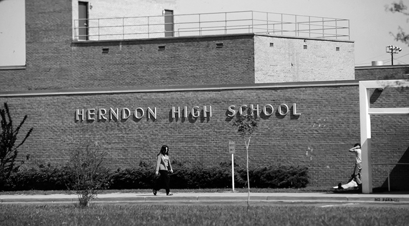 Black and white photograph of the front of Herndon High School’s 1967 building. The name Herndon High School is attached to the brick façade. One student can be seen walking along the sidewalk and two more can be seen on the far right side of the image.