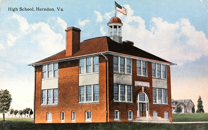 Color postcard of the first Herndon High School, circa 1920. The building is a square, two-story structure with brown brick walls and a brown tile roof. There is a cupola on the roof with an American flag atop it. There are two tall chimneys on either side of the building and large windows on every side. The main entrance, a set of double doors under a rounded archway, is reached by a paved walkway.